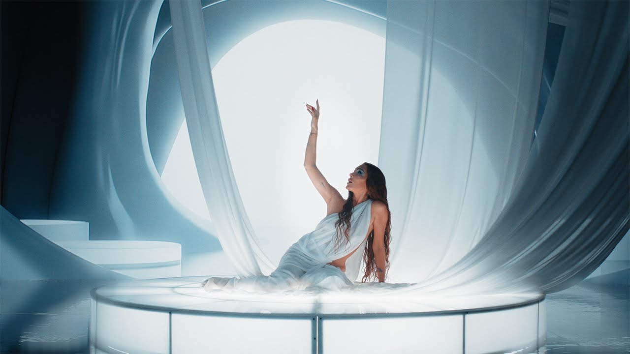 Woman in a flowing white dress sits on a circular lit platform, reaching upward among white fabric drapes in a futuristic scene.