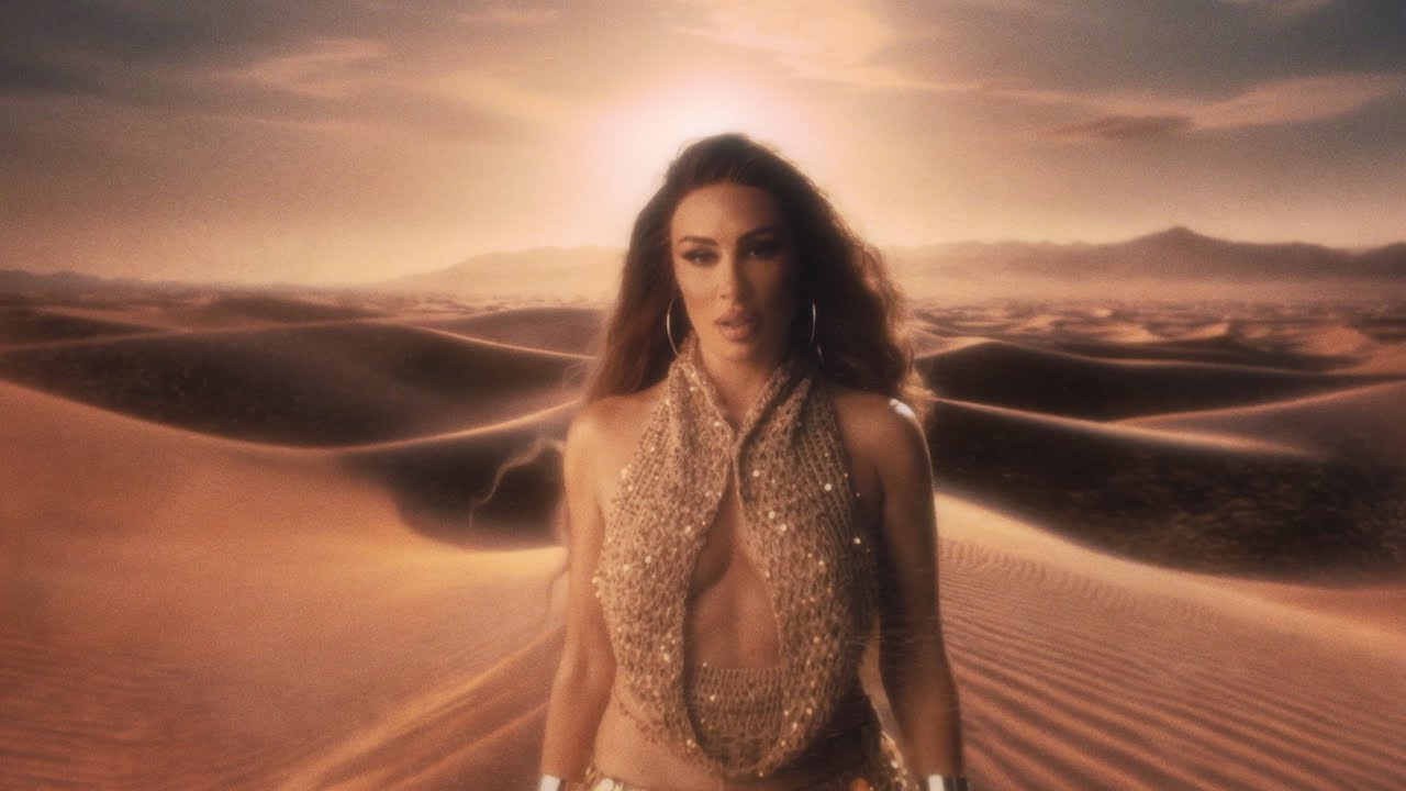Woman in a sparkling gold beaded halter top stands in desert dunes at sunset.